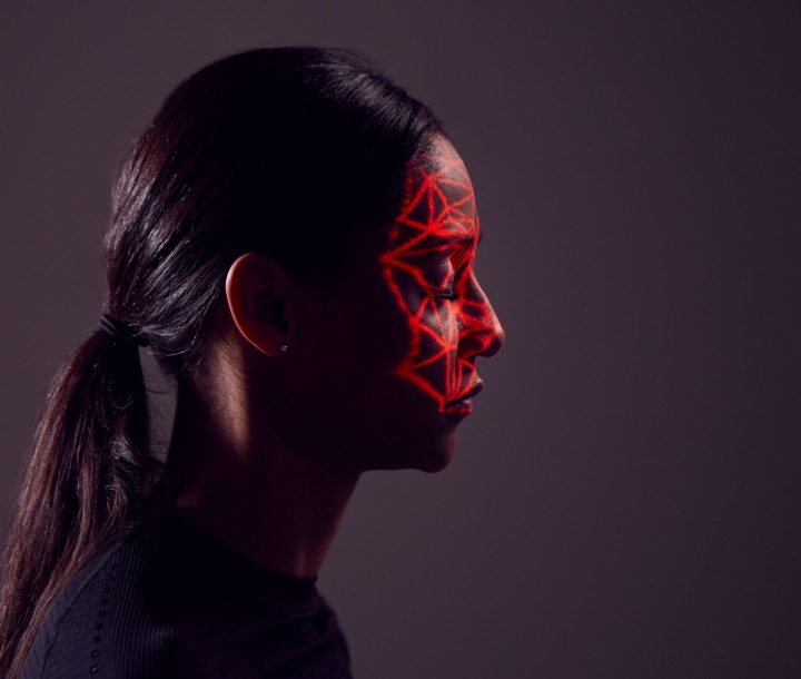 A women with a ponytail and black top has her face turned to the side, with a red light shining a web-like shadow on her face