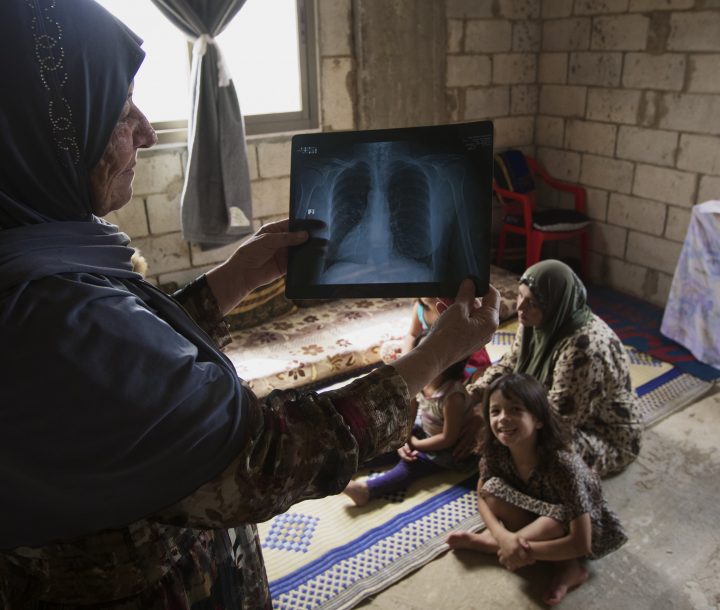 A woman wearing a headscarf holds up a chest Xray. A woman and two children sit on a rug on the ground.