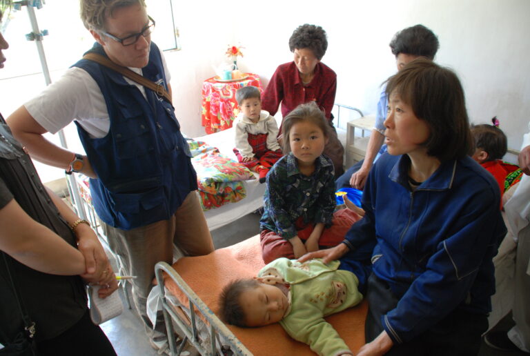 A woman sits on a hospital bed holding the hand of a small child. Another person in a blue vest faces the woman as a small child looks on