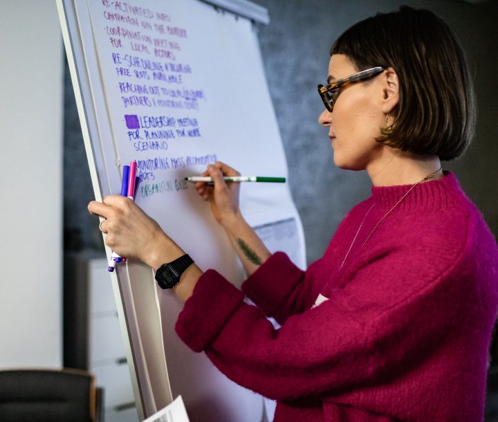 A woman wearing a pink top and glasses writes on a large piece of paper on a whiteboard.