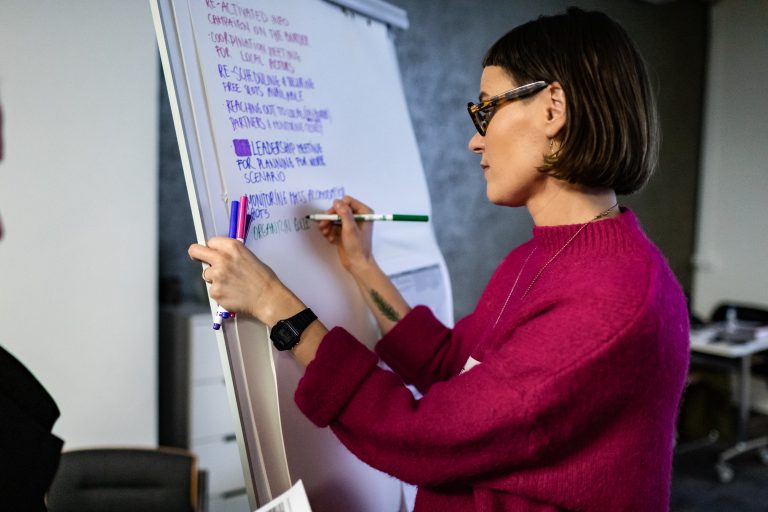 A woman wearing a pink top and glasses writes on a large piece of paper on a whiteboard.