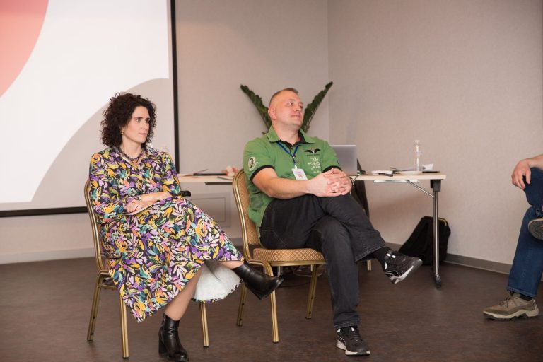 Two people sit on chairs facing forward with a projector screen behind them.