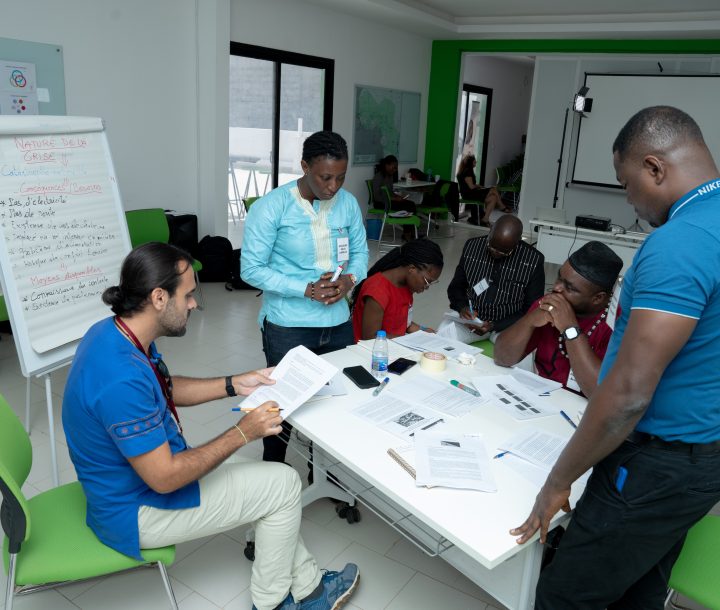 A group of five people are gathered around a table looking at pieces of paper. There is a note board to the left of the group.